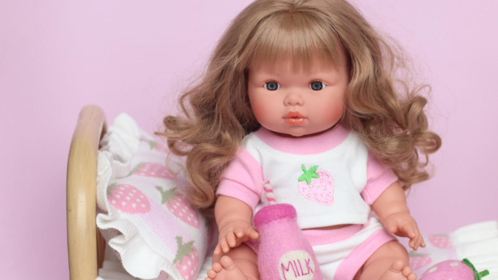 Mini Collettos llorens Doll in pink strawberry outfit sitting on a bed with strawberry patterned bedding against a pink background